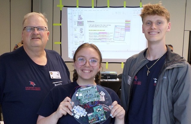 A young woman, flanked by two men, holds a dinner-plate-sized acrylic disk with computer boards attached to it